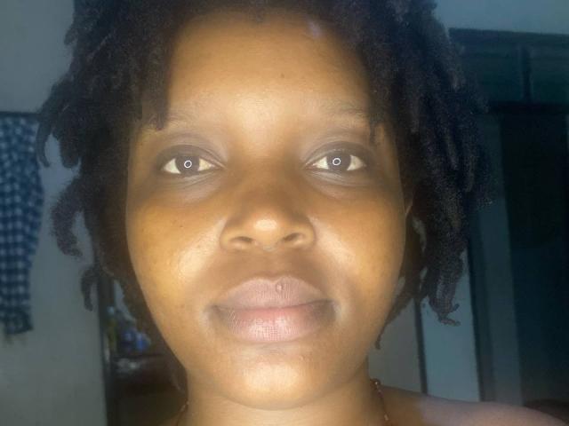 Close-up of SexyArya's face. She has dark brown skin, medium-sized lips with a slight upward curve, and small brown eyes. Her hair is short and curly, framing her face. She's wearing a simple gold necklace. The light source is directly in front, highlighting her face while the background is blurred. The overall composition is centered and focused on her facial features.