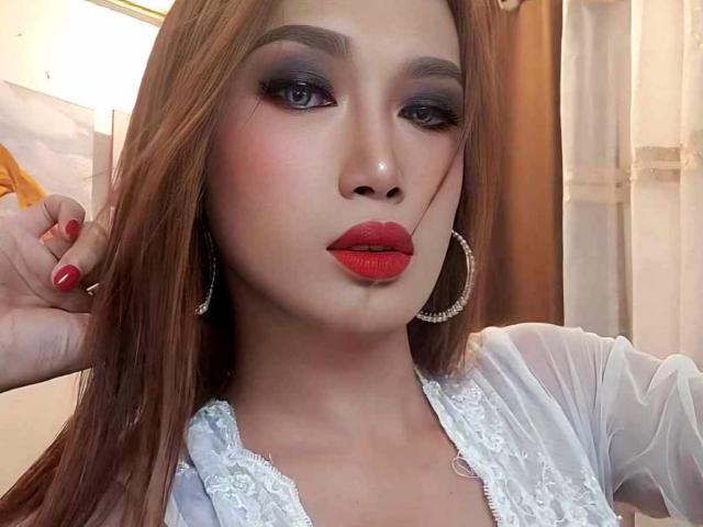 xGoddessJazzyx has red lipstick, dark eye makeup, and red nails, wearing a white lace top with hoop earrings, and long brown hair, looking at the camera.