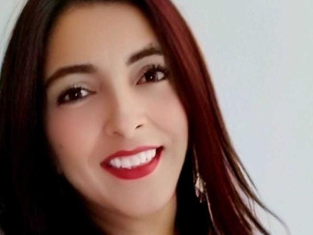 SamanthaXRossie with dark red hair, light skin, brown eyes, red lipstick, and a smile, wearing small earrings, in a headshot. Her tits are slightly visible under her collar.
