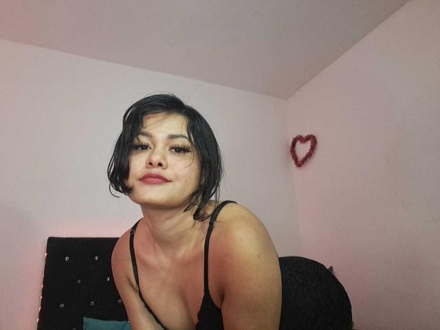 JennLunaLee with short black hair, wearing a black spaghetti-strap top, leaning forward, showing her tits, with a red heart wall decoration and a black headboard in the background.