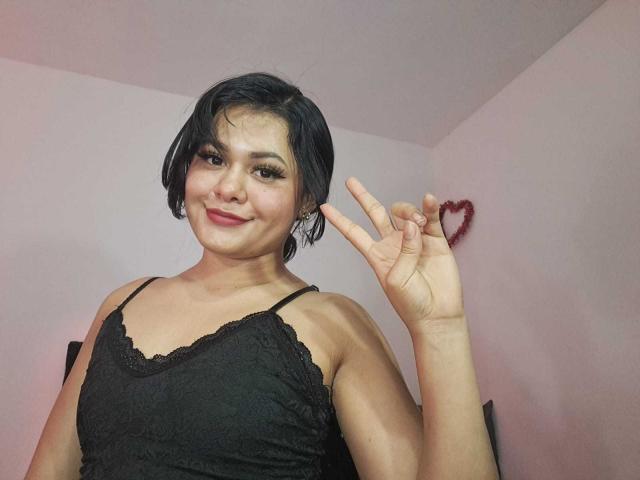 JennLunaLee with short black hair, smiling, wearing a black lace camisole, making a peace sign with her right hand, red heart necklace.