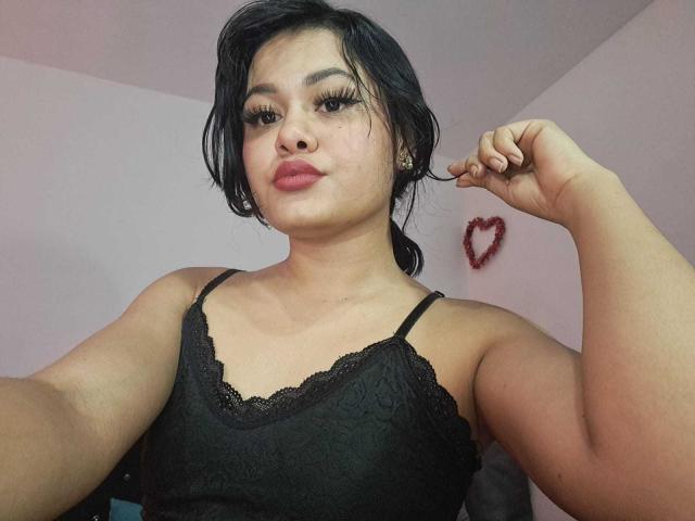 JennLunaLee with medium tits, wearing a black lace camisole, has dark hair in a messy bun, thick eyelashes, and a red heart tattoo on her right cheek.