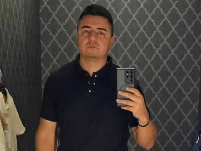 MisterMiggie stands in front of a mirror, holding a smartphone in his right hand. He has short, dark hair and a neutral expression. He wears a black, short-sleeved, button-up shirt. The background features a patterned, dark grey wall. The image is a selfie, with MisterMiggie's face centered and his upper body visible. The smartphone is black, matching the wall pattern. The lighting is even, highlighting MisterMiggie's facial features.