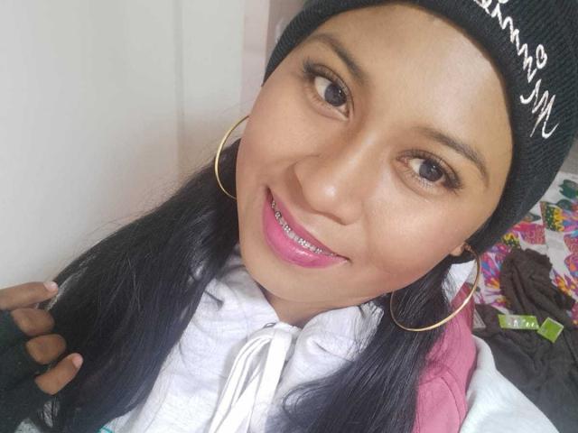 EbonyxLust, con piel morena, gorro negro, aros de oro, lápiz labial rosa y cabello negro lacio, sonriendo a la cámara, vistiendo una sudadera blanca..