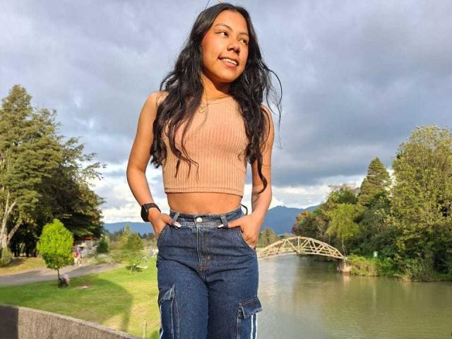 IvyMontaner with long black hair, brown crop top, and blue jeans, hands in pockets, standing by a river, overcast sky, smiling, wearing a black smartwatch.
