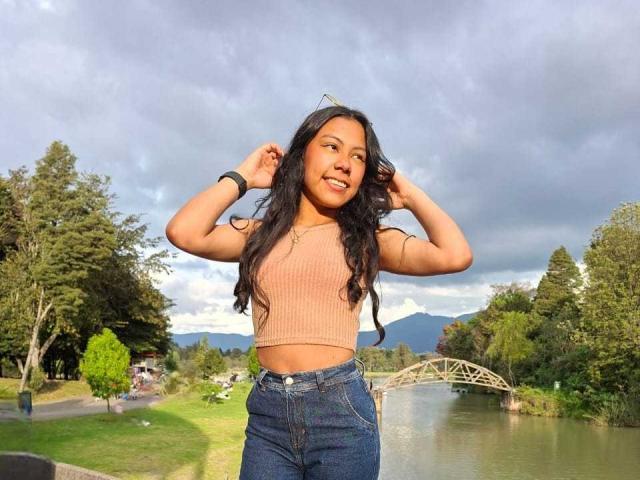IvyMontaner with long black hair, tan crop top, and blue jeans, standing by a river, smiling, hands up, wearing a black bracelet, sunny, cloudy sky, green park.