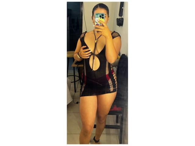Velka, wearing a black, sheer, cut-out dress with red stripes, showing her tits and pussy through the fabric, taking a mirror selfie with a phone.