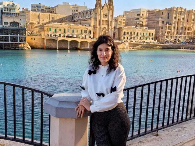 AddictedToUTS stands in front of a black metal railing, leaning on a gray concrete post. She has curly dark hair, a light skin tone, and wears a white long-sleeve shirt with black patterns, and black textured pants. She's smiling slightly, looking at the camera. The background features a blue-green waterway with a historic stone building and clock tower. The image is clear with natural light.