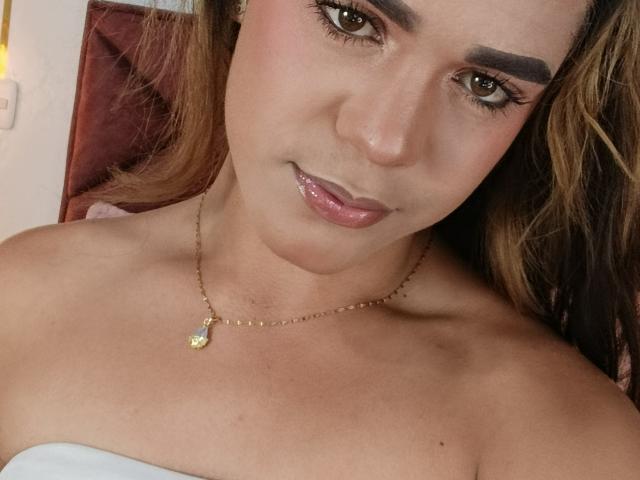AliceCoolenk with light brown skin, long wavy brown hair, thick eyebrows, brown eyes, glossy pink lips, gold necklace, and bare shoulders, smiling softly at the camera.