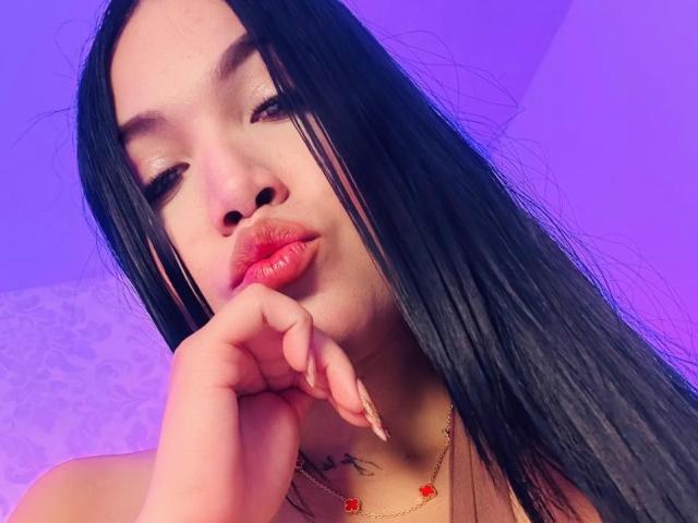 MiaDenver, com longos cabelos pretos, lábios vermelhos carnudos e um piercing no nariz, tocando o queixo com o dedo, usando um colar de ouro com flores vermelhas, sob iluminação roxa..