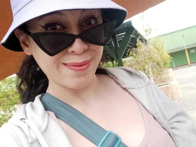 KendraSimons with black sunglasses, pink lipstick, grey hoodie, and teal seatbelt, wearing a purple bucket hat, showing her tits slightly through a pink ribbed top.