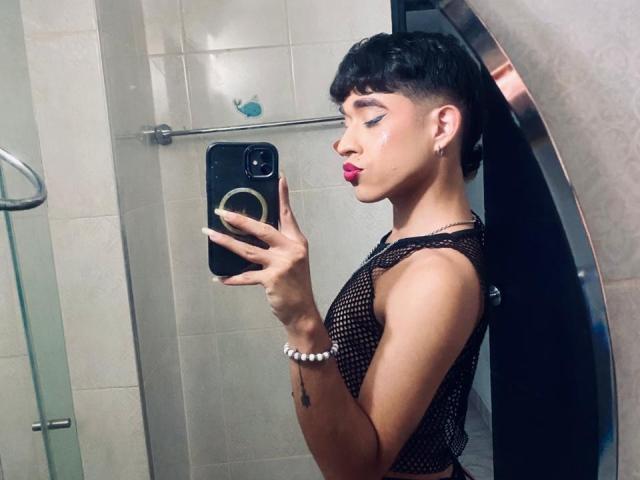 BriggitteLove with short black hair, pink lipstick, fishnet top, holding phone in bathroom mirror, showing her tits, bracelet on wrist, tattoo on arm, looking at phone.