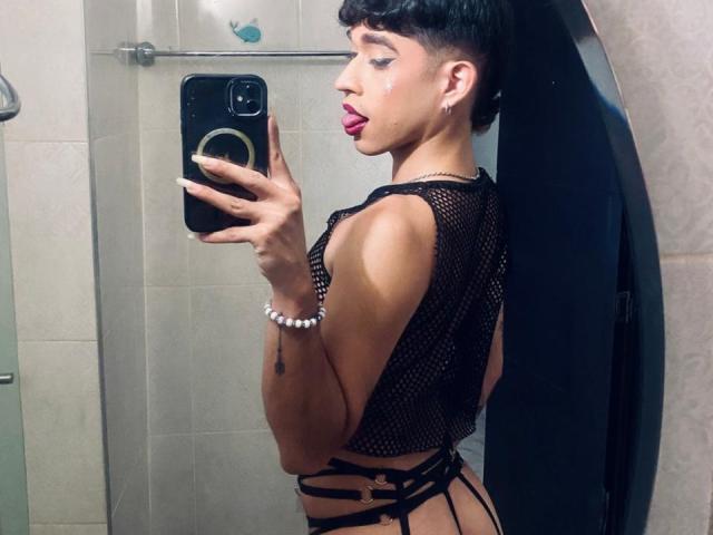 BriggitteLove with short black hair, pink lipstick, and a mesh top, showing her side with strappy black underwear, holding a phone, in a mirror selfie.