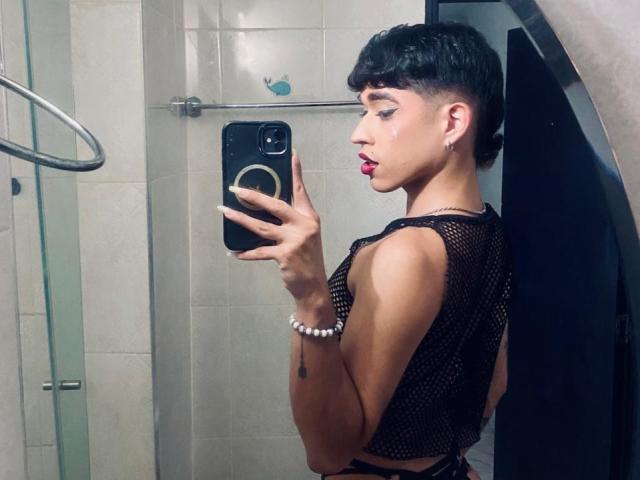 BriggitteLove, a young person with a dark short haircut and bold red lipstick, holds a black phone with a ring strap in a bathroom. They wear a black mesh crop top, exposing their back, and a white bead bracelet. Their right arm has a small tattoo. The bathroom has white tiled walls and a glass shower door on the left. The image is a selfie.