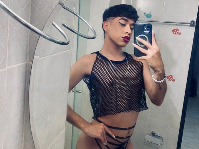 BriggitteLove with short brown hair, red lipstick, fishnet top, black strappy panties, holding phone in mirror selfie, showing slim, toned body and small tits. Bathroom setting.