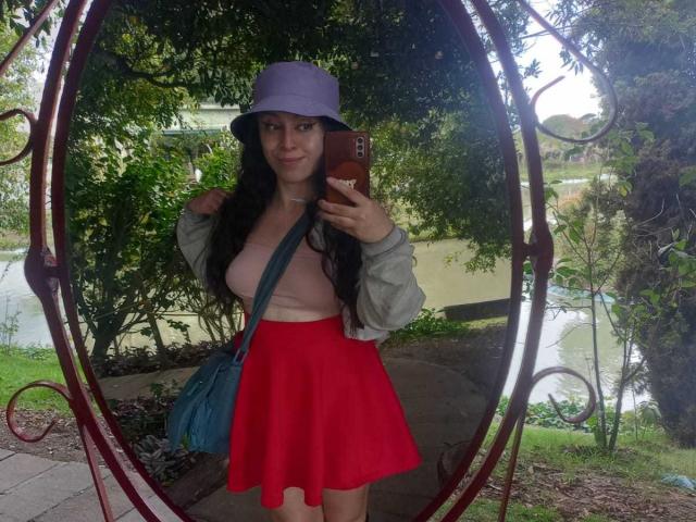 KendraSimons, con cabello largo y negro, blusa rosa, falda roja, chaqueta gris, gorro tipo cubo morado, bolso azul, sosteniendo un teléfono, selfie frente a un espejo, al aire libre, vegetación, agua en el fondo, sonrisa segura..