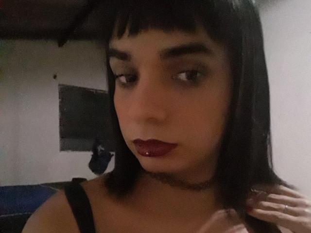 Aripietra with dark brown bob, black choker, red lipstick, and black strap dress, looking down, hand near chin, soft light, slight shadow on face, brown eyes, subtle makeup.