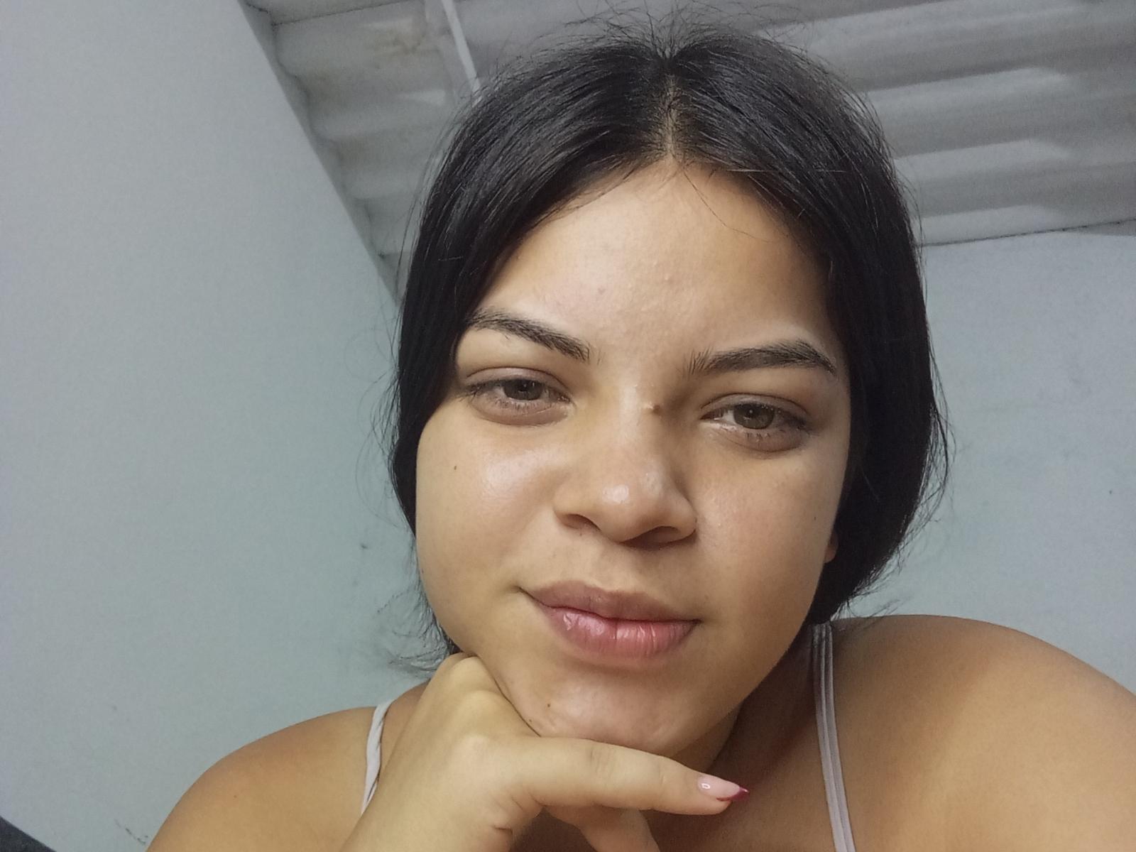 TeresaFrancie has dark hair, brown eyes, and a slight smile with her finger on her chin. She's wearing a gray tank top, showing her bare shoulders and chest.