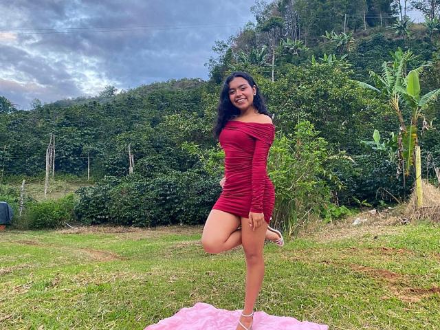 JuliaBennett standing on grass, wearing a red off-shoulder dress, holding one leg up, smiling, with black hair, pink shoes, and a pink cloth on the ground.