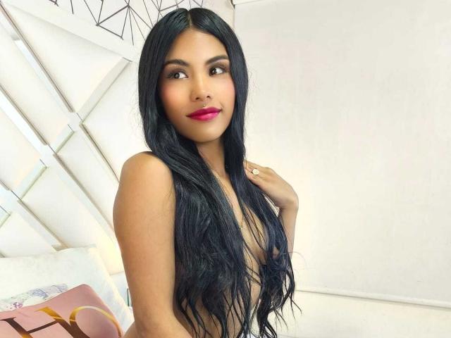 ClarisCampbell has long black hair, pink lipstick, and brown eyes. She's topless with her hand on her shoulder, showing her small tits. She's smiling slightly at the camera.