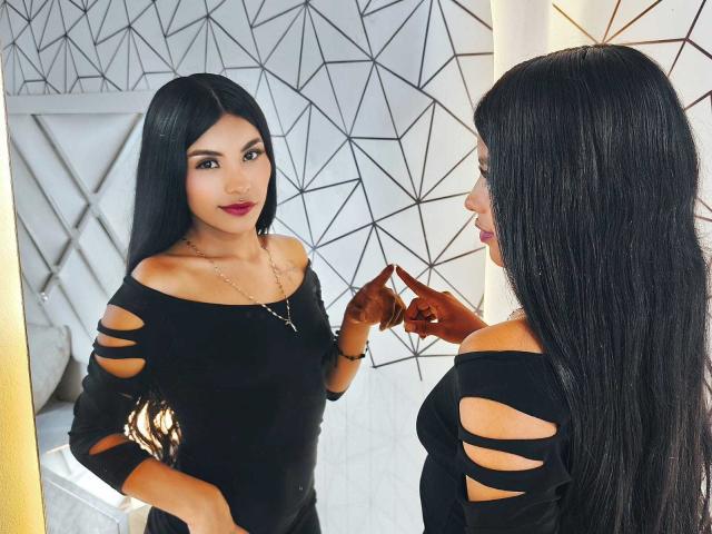 ClarisCampbell with long black hair, wearing an off-shoulder black dress with cut-out sleeves, standing in front of a mirror, touching her nose, showing her smooth, tan skin.