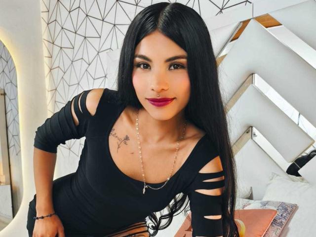 ClarisCampbell with long black hair, wearing a black cut-out dress, showing small tits, tattoos on chest, necklace, red lipstick, leaning forward, smiling.