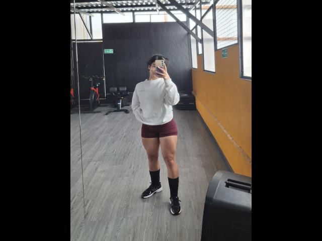 PaulethJoens with a white hoodie, maroon booty shorts, black socks, and black sneakers, standing in a gym, taking a mirror selfie, showing her toned ass and legs.
