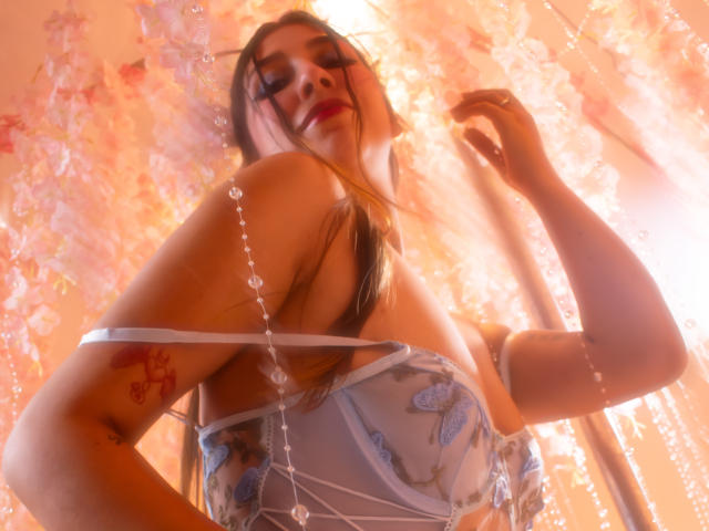 BellyRouse with long brown hair, red lipstick, and blue floral bra, beads hanging, tattoo on left arm, low angle, glowing light, sexy, confident, soft focus, warm colors.