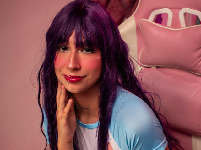 BellyRouse with purple hair, pink blush, and shiny pink lipstick, wearing a blue and white shirt, touching her cheek, sitting in front of a pink chair.