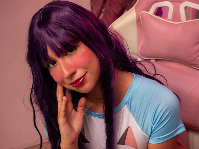 BellyRouse with purple hair, pink blush, and red lipstick, wearing a blue and white raglan shirt, touching her cheek, smiling softly. Her hand is on the right side.