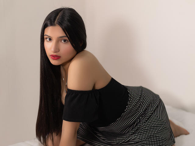 LaureneLove with long black hair, red lips, off-shoulder black top, and black polka dot skirt, kneeling on a white surface, looking at the camera.