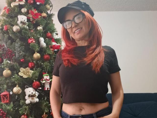 MarianaMonsalve with red hair, black cap, glasses, black crop top, showing her navel, standing in front of a decorated Christmas tree, smiling, wearing blue jeans.