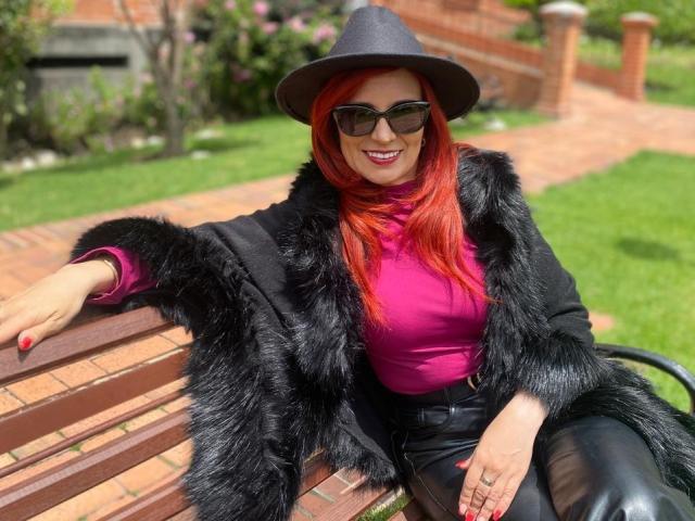 MarianaMonsalve with red hair, black fur coat, purple top, black pants, black hat, and sunglasses, sitting on a brick bench, smiling, showing her ass with leather pants.