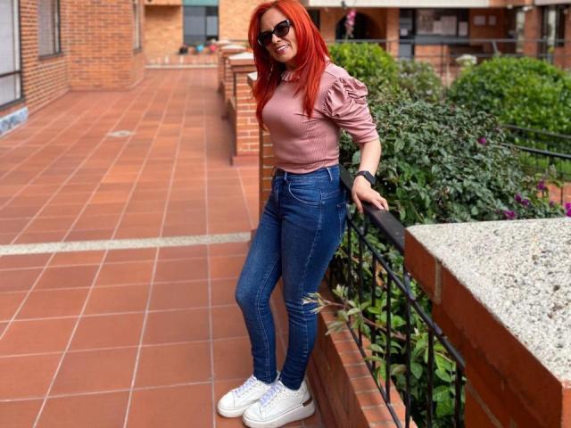 MarianaMonsalve with red hair, wearing sunglasses, pink top, blue ripped jeans, white sneakers, leaning on black railing, smiling, brick balcony, green plants, red tiles.