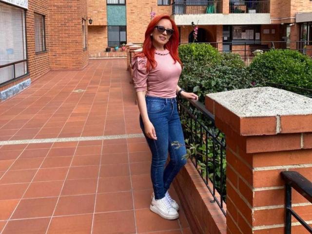 MarianaMonsalve has red hair, wears a pink top, blue ripped jeans, white sneakers, and sunglasses. She stands on a brick balcony, leaning on a black railing.