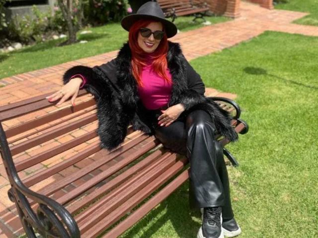 MarianaMonsalve with red hair, black hat, sunglasses, black fur coat, pink top, black leather pants, black shoes, sitting on wooden bench, green grass, brick path, smiling.