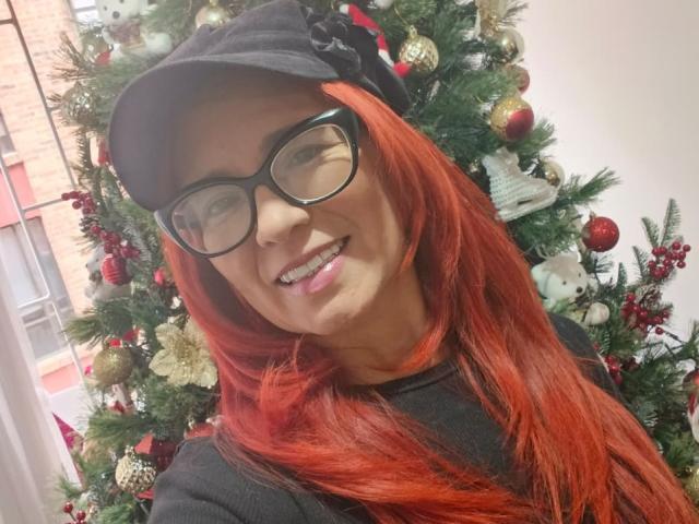 MarianaMonsalve with red hair, black glasses, and black cap, smiling, showing her teeth, standing in front of a decorated Christmas tree with red and gold ornaments.