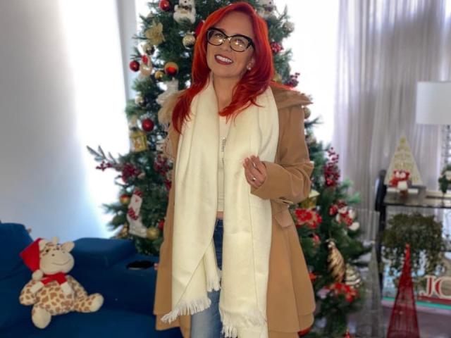 MarianaMonsalve with red hair, glasses, tan coat, white scarf, smiling, standing in front of decorated Christmas tree, blue couch with giraffe toy, and white curtains.