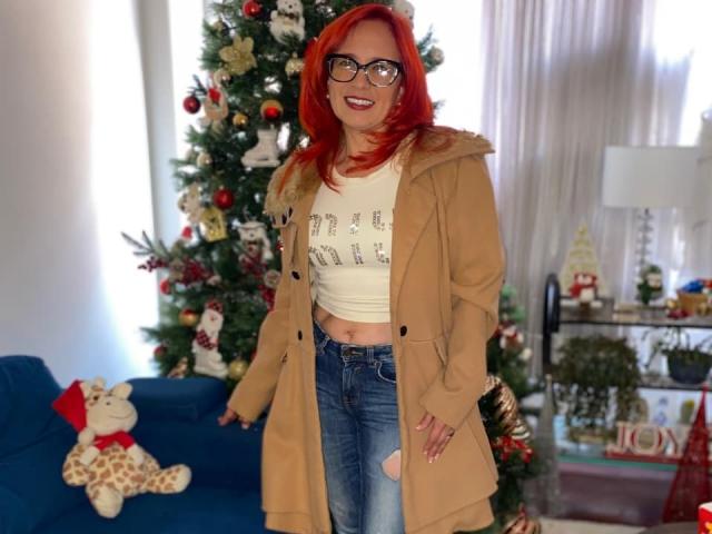 MarianaMonsalve with red hair, glasses, smiling, tan coat, white shirt, "HOLIDAY" text, blue jeans, standing by Christmas tree, blue sofa, giraffe toy, and white curtains.