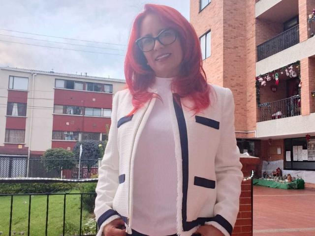MarianaMonsalve with red hair, glasses, white jacket with black trim, and white top, standing outside, smiling, hands in pockets, looking cute and sexy.