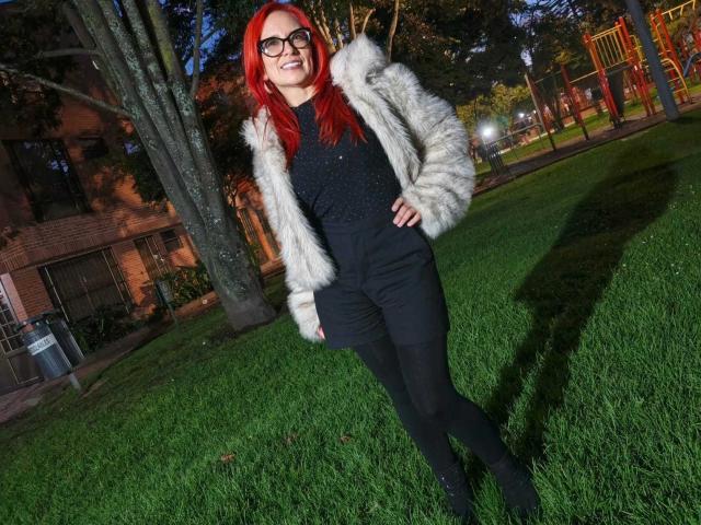 MarianaMonsalve with red hair, glasses, black top, black pants, black shoes, and white furry jacket, standing on grass, smiling, hands on hips, nighttime, park setting.