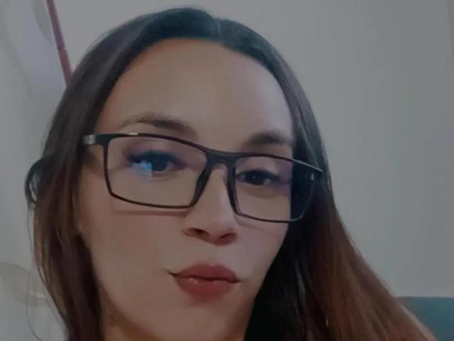 MelissaBxd, com lábios carnudos, usando óculos pretos, tem cabelo castanho e pele clara, olhando ligeiramente para o lado. Suas nádegas não estão visíveis..