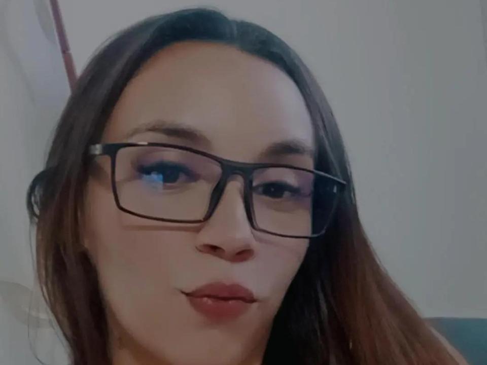 MelissaBxd, com lábios carnudos, usando óculos pretos, tem cabelo castanho e pele clara, olhando ligeiramente para o lado. Suas nádegas não estão visíveis..