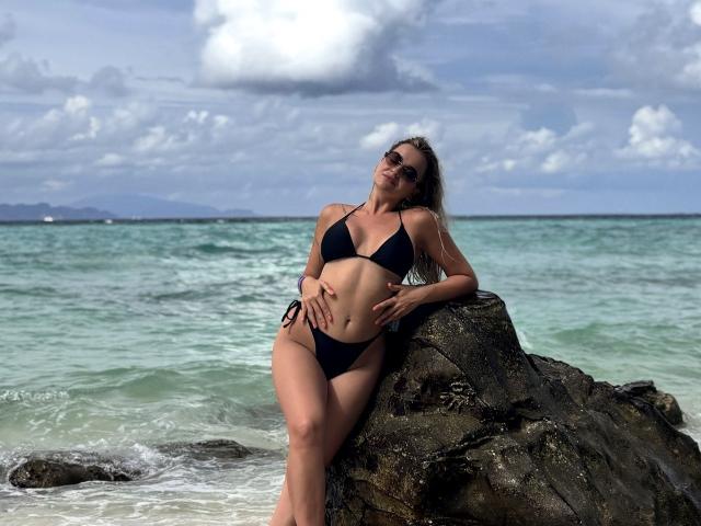 EvaAdderiy leans against a rock, wearing a black bikini. Her long hair is loose, and she wears sunglasses. She has a hand on her belly and the other on the rock. She's smiling. The rock is textured, dark, and wet from the ocean. The ocean is greenish-blue with small waves. The sky has fluffy clouds. EvaAdderiy's skin is tan, and she's slightly pregnant. The image is bright and sunny.