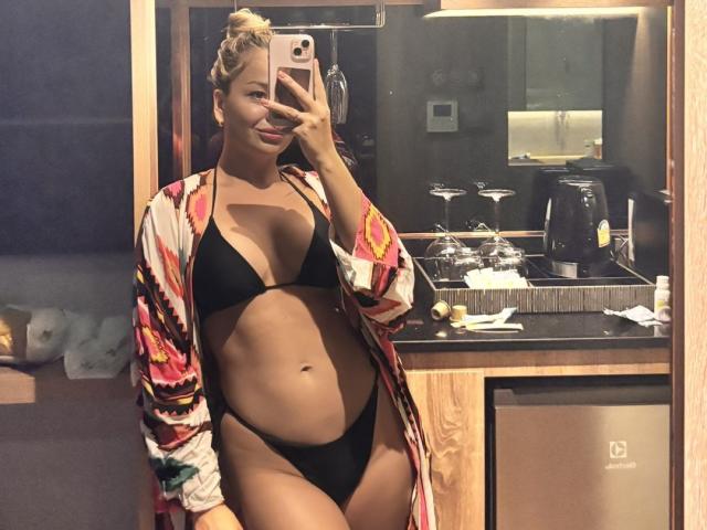 EvaAdderiy takes a mirror selfie in a bathroom. She has light brown skin and blonde hair in a bun. She wears a black string bikini with high-cut sides, revealing her belly and a slight curve. Over the bikini, she has on a colorful, patterned cardigan. She holds a pink smartphone in her right hand. The background includes a black countertop with a coffee maker, glasses, and a trash can. The image has a casual, intimate vibe. The lighting is warm and slightly dim.