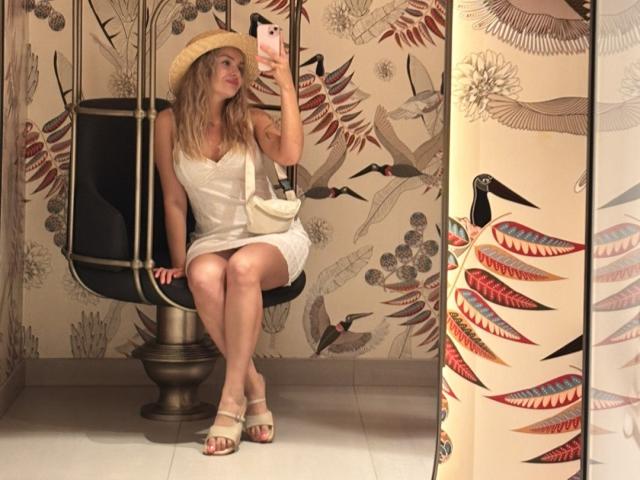 EvaAdderiy sitting on a black swing, wearing a white dress, straw hat, and sandals, holding a phone, with a beige bag on her shoulder, in a patterned room.
