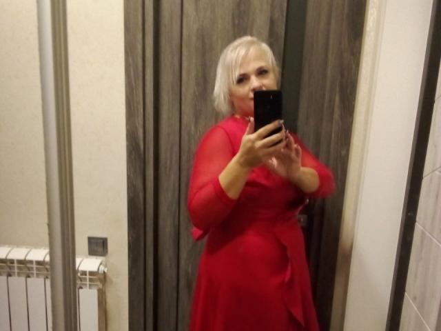 CrystalVibe stands in a mirror selfie, holding a black smartphone. She has short, platinum blonde hair and wears a bright red, sheer dress with long sleeves. Her right hand holds the phone, obscuring part of her face. The dress's texture is slightly translucent, revealing her skin tone underneath. The background is a wooden door. The lighting is even, highlighting her dress's color and texture. The image is candid and focused on her reflection.