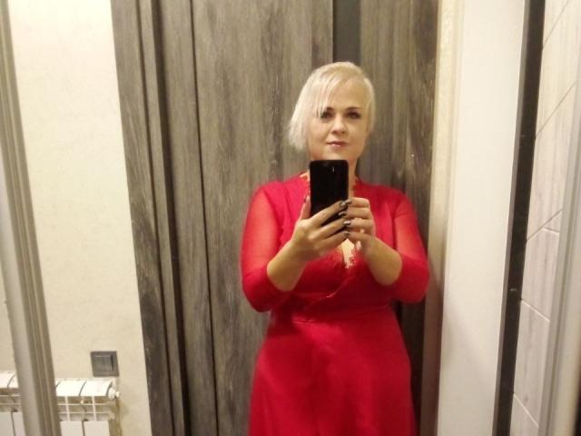 A selfie of CrystalVibe with platinum blonde hair, wearing a sheer red dress with a lace neckline. She holds a black phone in her right hand, standing in a room with wooden doors and white tiled walls. Her expression is neutral, and she looks directly at the camera. The room has a radiator on the left and a grey electrical outlet near the bottom left corner. The texture of the dress is shiny and transparent.