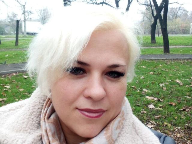 CrystalVibe has short, messy white hair and fair skin. She has dark eyeliner and pink lipstick. She's wearing a beige, fluffy scarf and a dark jacket. The image focuses on her face, which is centered, with a slight smile. The background is a grassy park with scattered leaves and trees. The photo has a natural, slightly overexposed look.