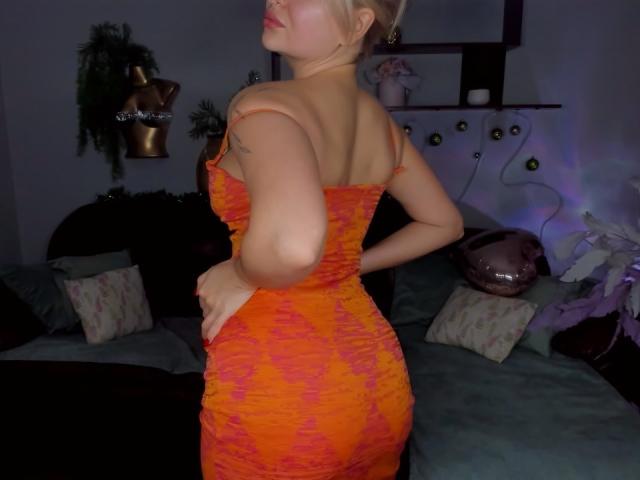 EvaAdderiy in an orange, ruched dress, standing with her back to the camera, showcasing her ass and a tattoo on her right arm. Her blonde hair is in a bun.